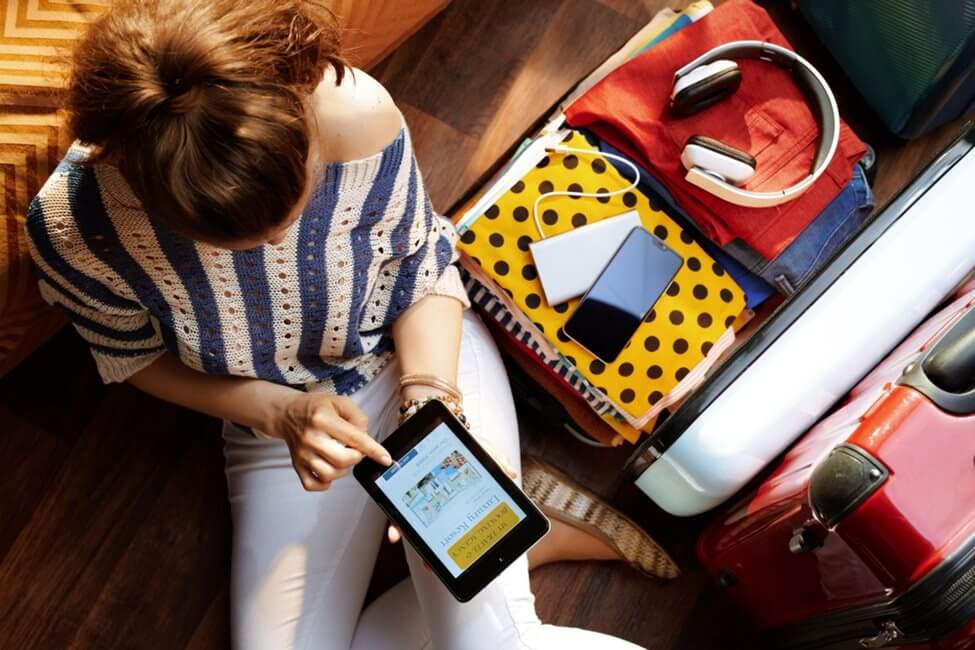 Birds eye view of a woman on her tablet, sat cross legged next to an open suitcase with a pair of headphones on top of clothes.