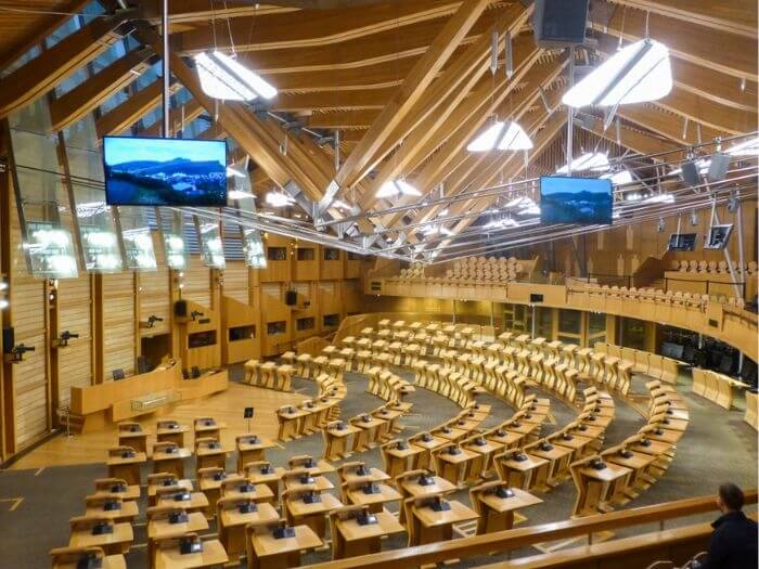 Inside of the Scottish Parliament at Holyrood, empty wooden seats sit in a half circle around the main speaking stage with screens.