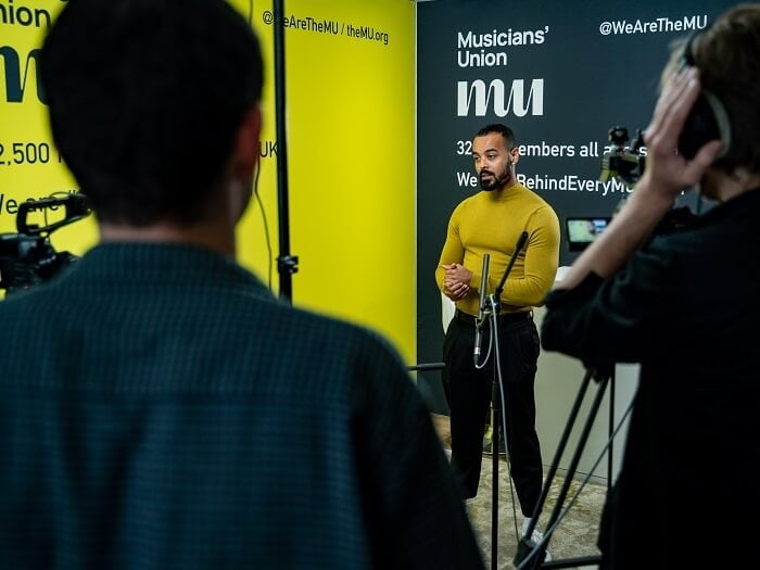 Linton Stephens talking into a microphone on a stand in front of a background of Musicians' Union banners.