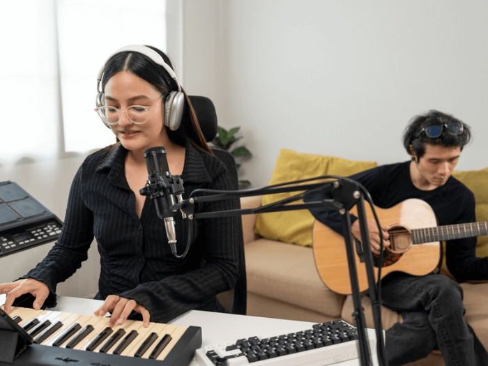 Young women playing keyboard in front of a mic and pop shield in a home studio. Behind her is a young man playing acoustic guitar.