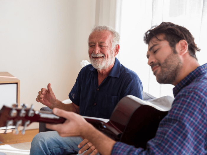 A young male musician, playing to an older man who is smiling as he listens.
