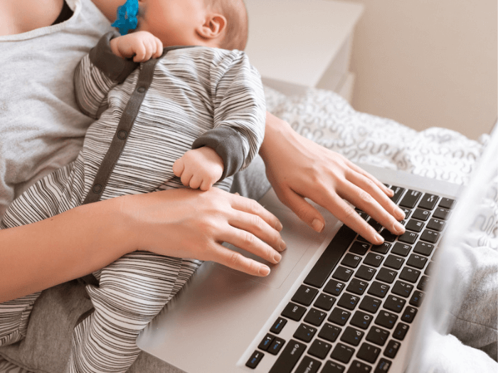 Mother holding a young baby while she's working on a laptop.