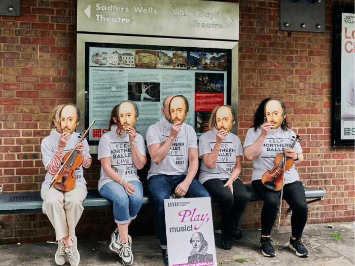 Five orchestra members sat at a bus stop with ‘Keep Northern Ballet Live’ t-shirts and wearing Shakepeare masks as part of our demo in May last year.
